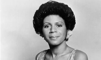 Black-and-white studio portrait of a woman with a short curly hairstyle, wearing a sleeveless top and earrings, smiling softly