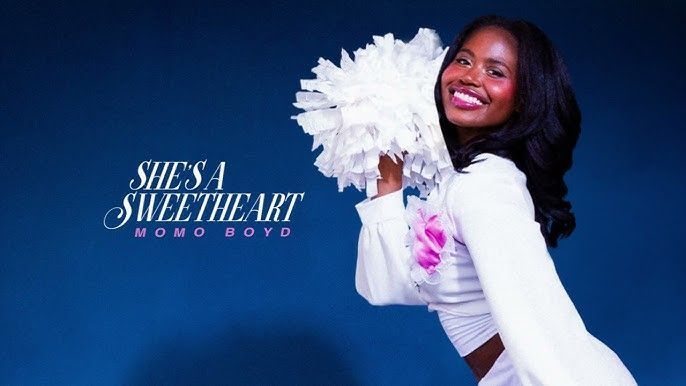 Woman smiling in a white outfit with white pom-poms against a blue background, text reads “SHE’S A SWEETHEART”