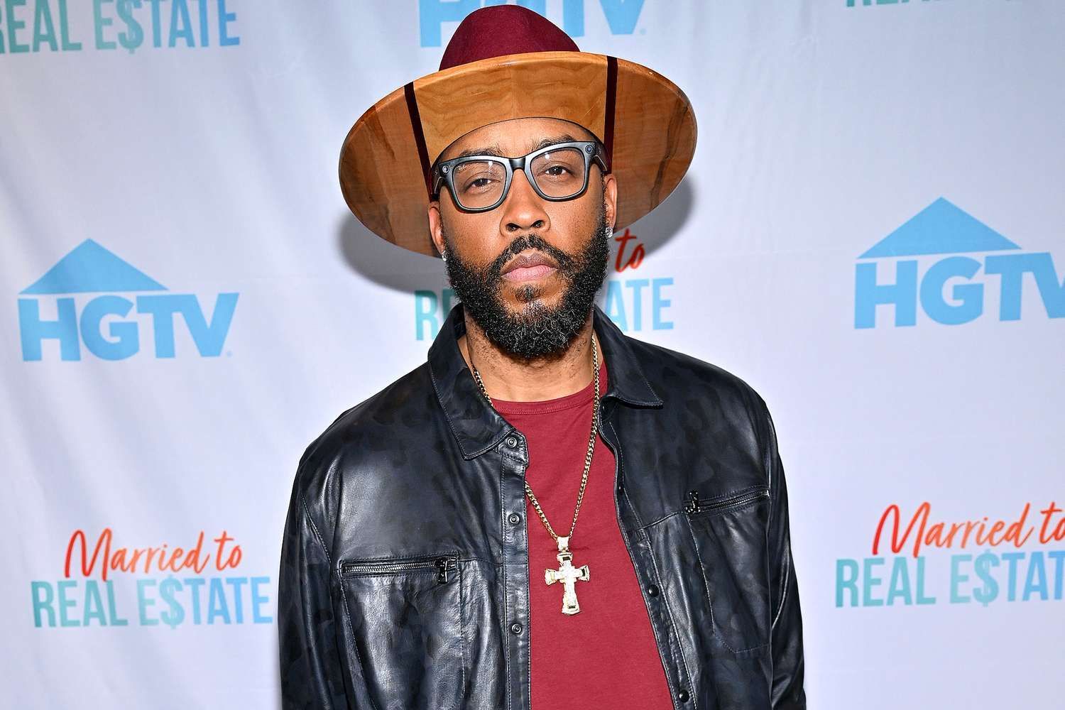Man in hat and glasses posing on red carpet before HGTV Married to Real Estate backdrop