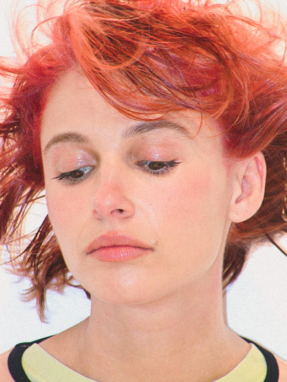 Close-up portrait of a person with short red hair and light makeup, looking downward against a pale background
