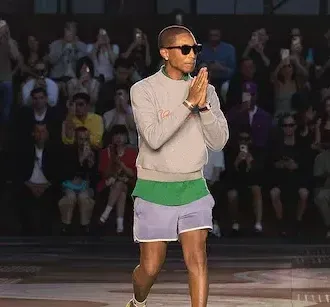 Person in sunglasses and athletic clothes claps while walking on a runway, with an applauding audience behind.