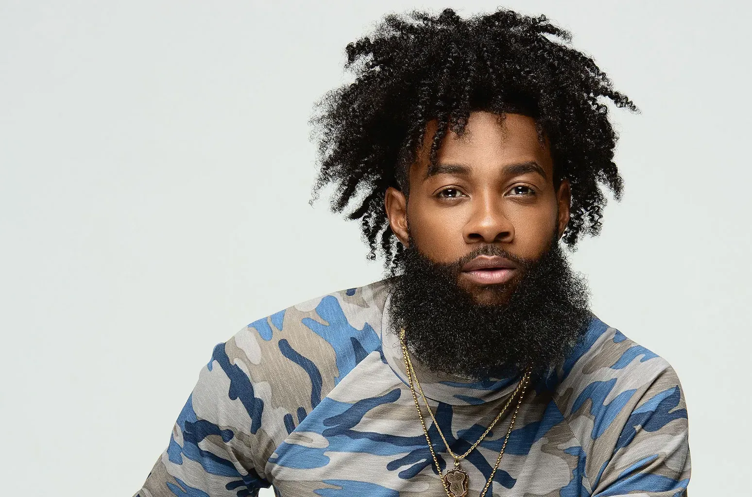 Man with curly hair and beard in a camouflage shirt, facing forward against a light gray background