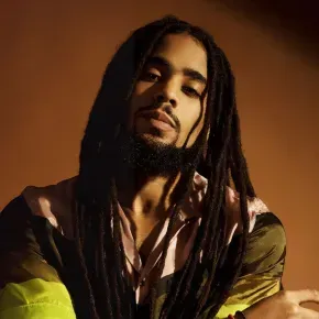 Portrait of a person with long dreadlocks against a warm brown background, wearing a green and pink top