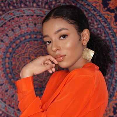 Woman in a bright orange top posing in front of a patterned tapestry, resting her chin on her hand.