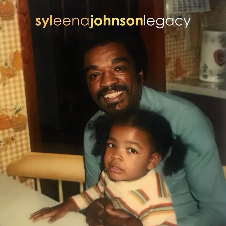 Man smiling behind a seated child in a kitchen, with warm lighting and a tiled background.