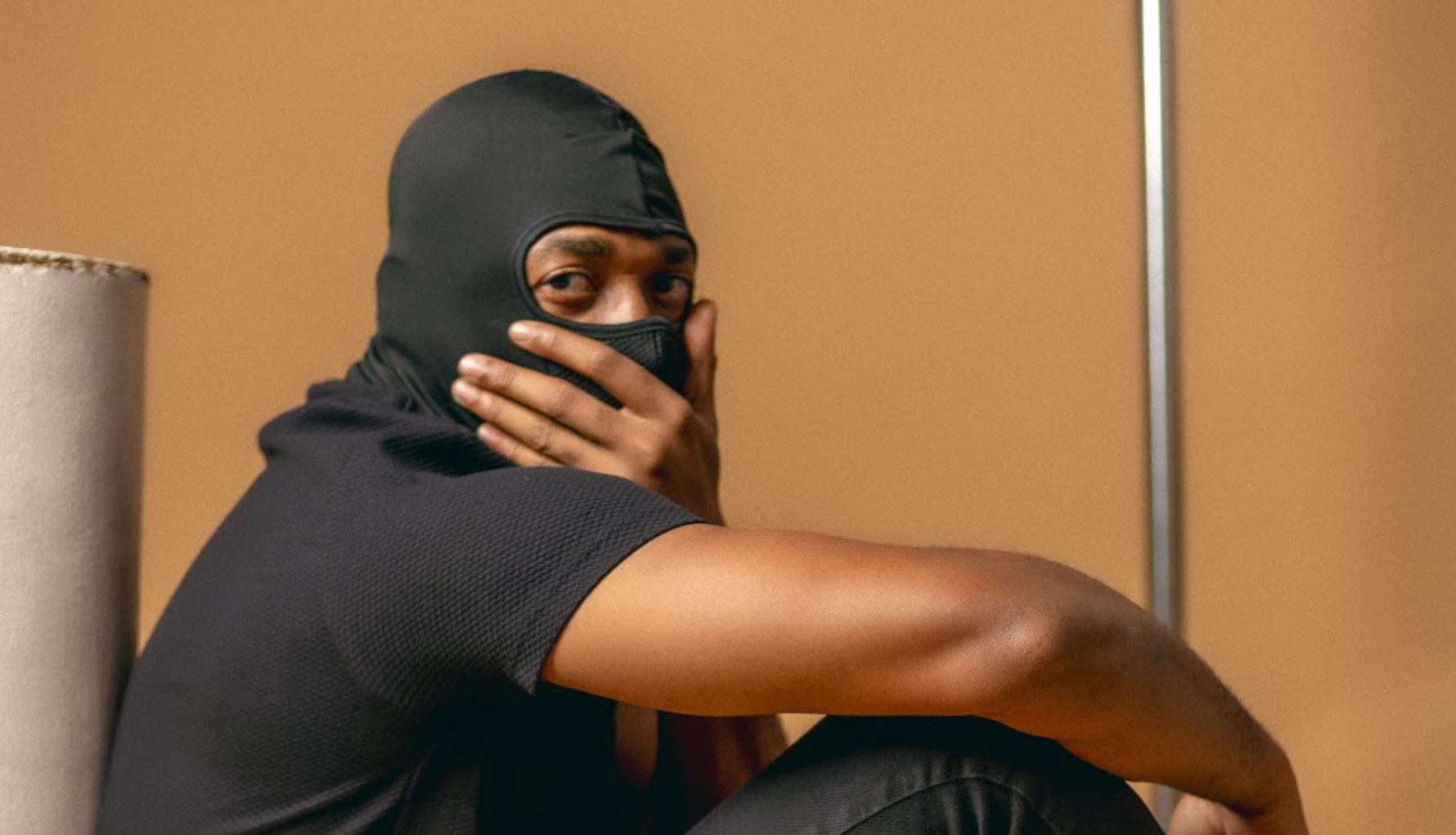 Person in a black balaclava covers their mouth while seated against a tan wall.
