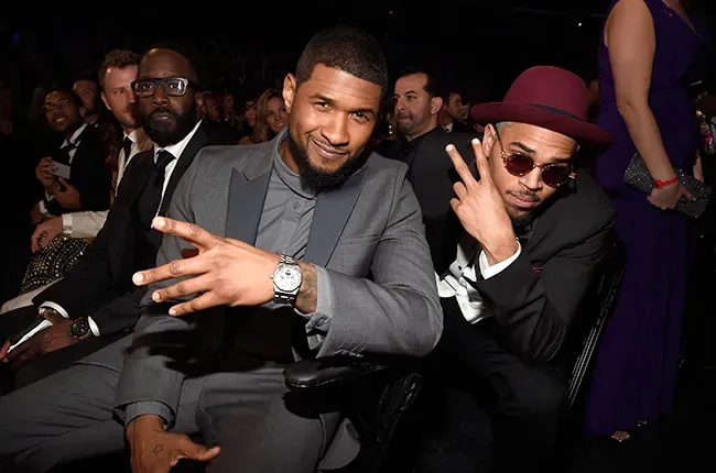 Two men posing at a formal event, one in a gray suit smiling and one in a burgundy hat and sunglasses making a hand sign.
