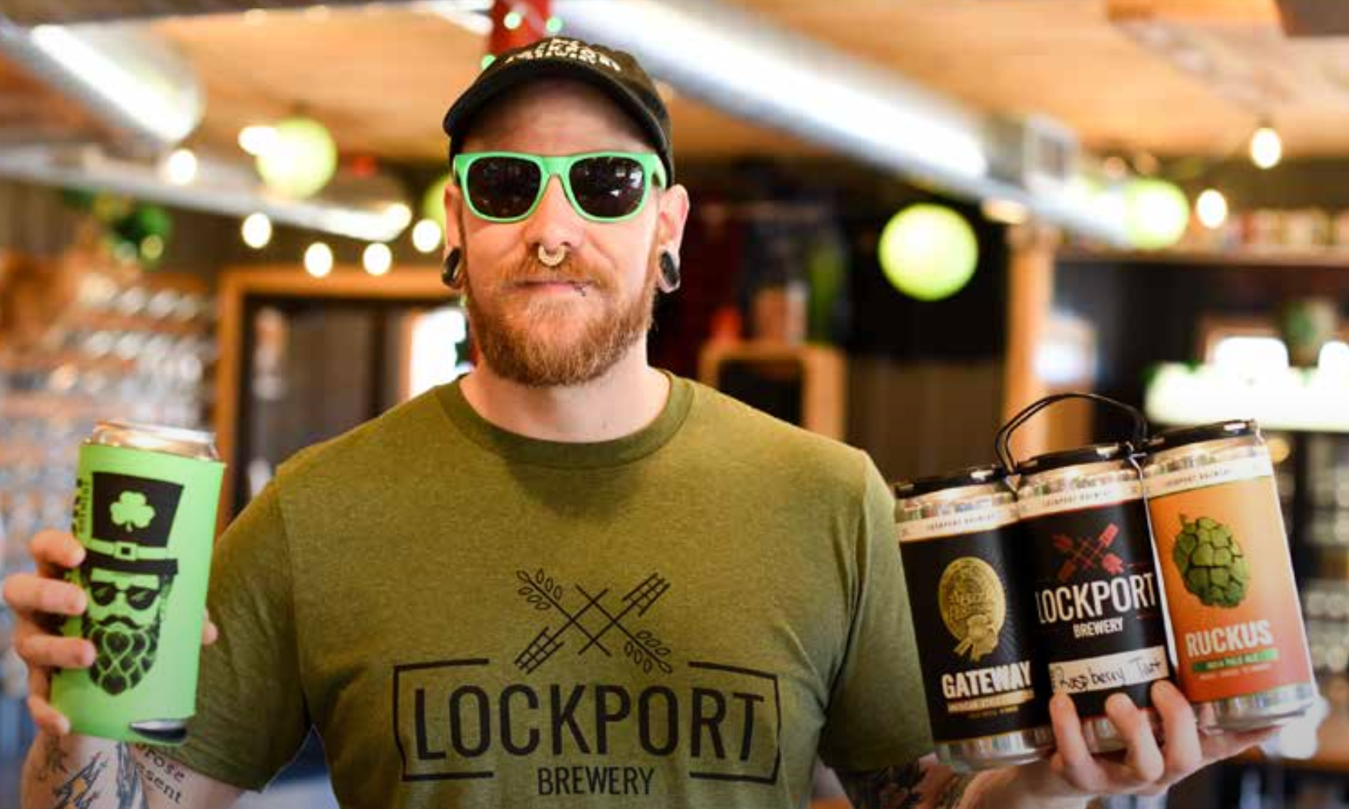 A man wearing sunglasses and a lockport brewery shirt is holding four cans of beer.