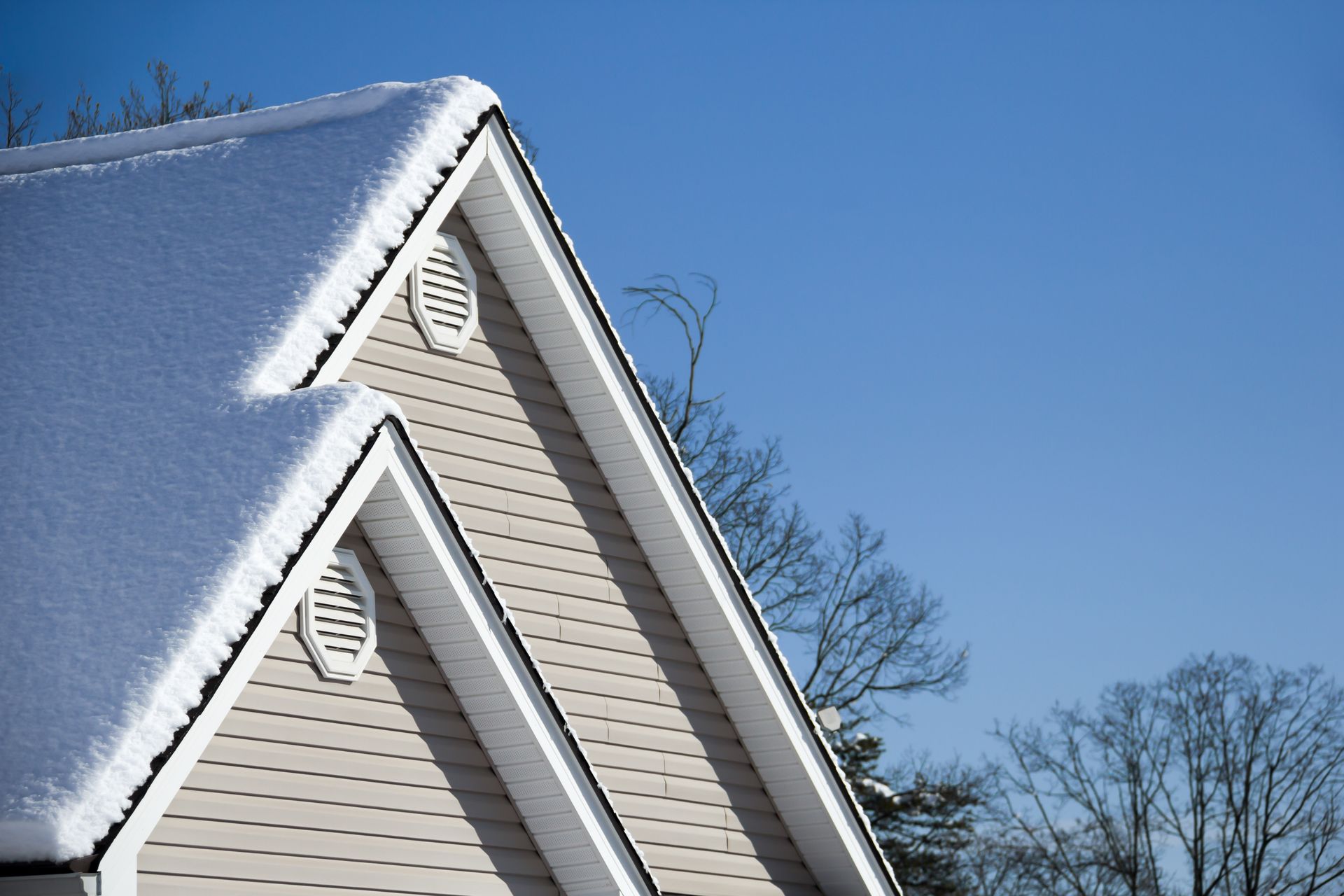 Un toit à pignon enneigé sur une maison beige se détachant sur un ciel bleu clair, avec deux aérations rondes au niveau du pignon.