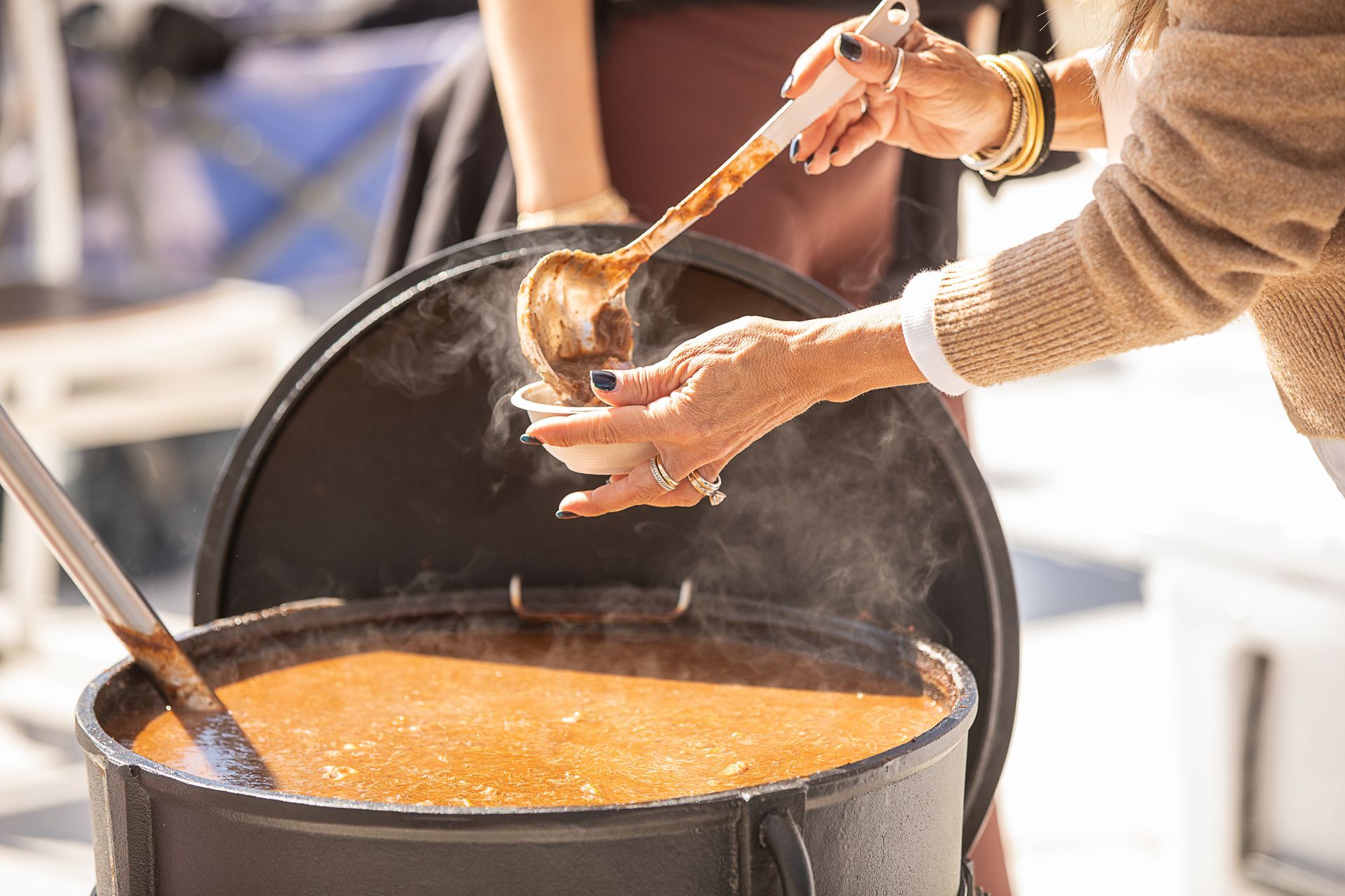 Hands ladling gumbo from a large black pot outdoors