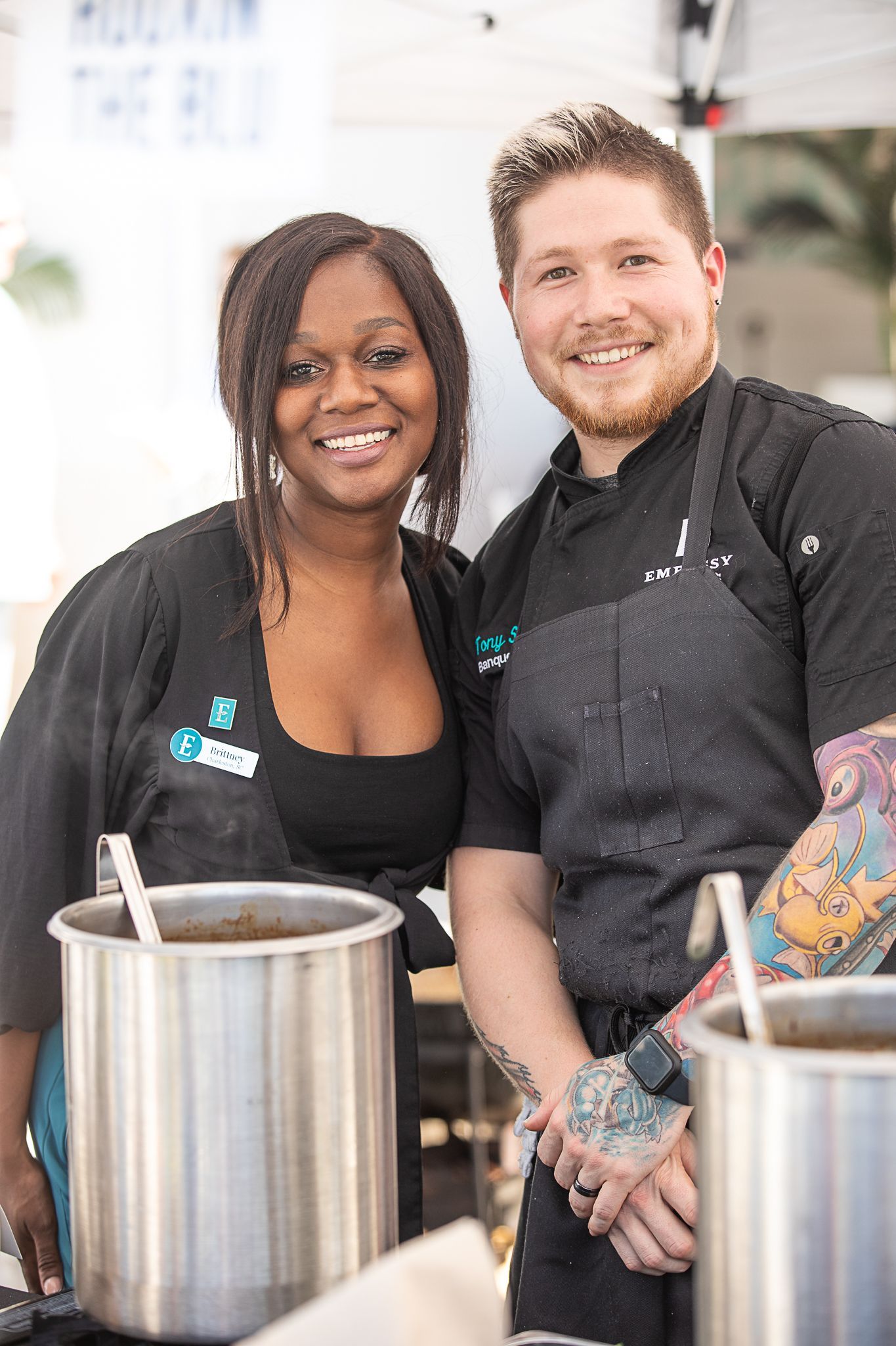 Two smiling chefs in black aprons standing behind large pots in a kitchen.