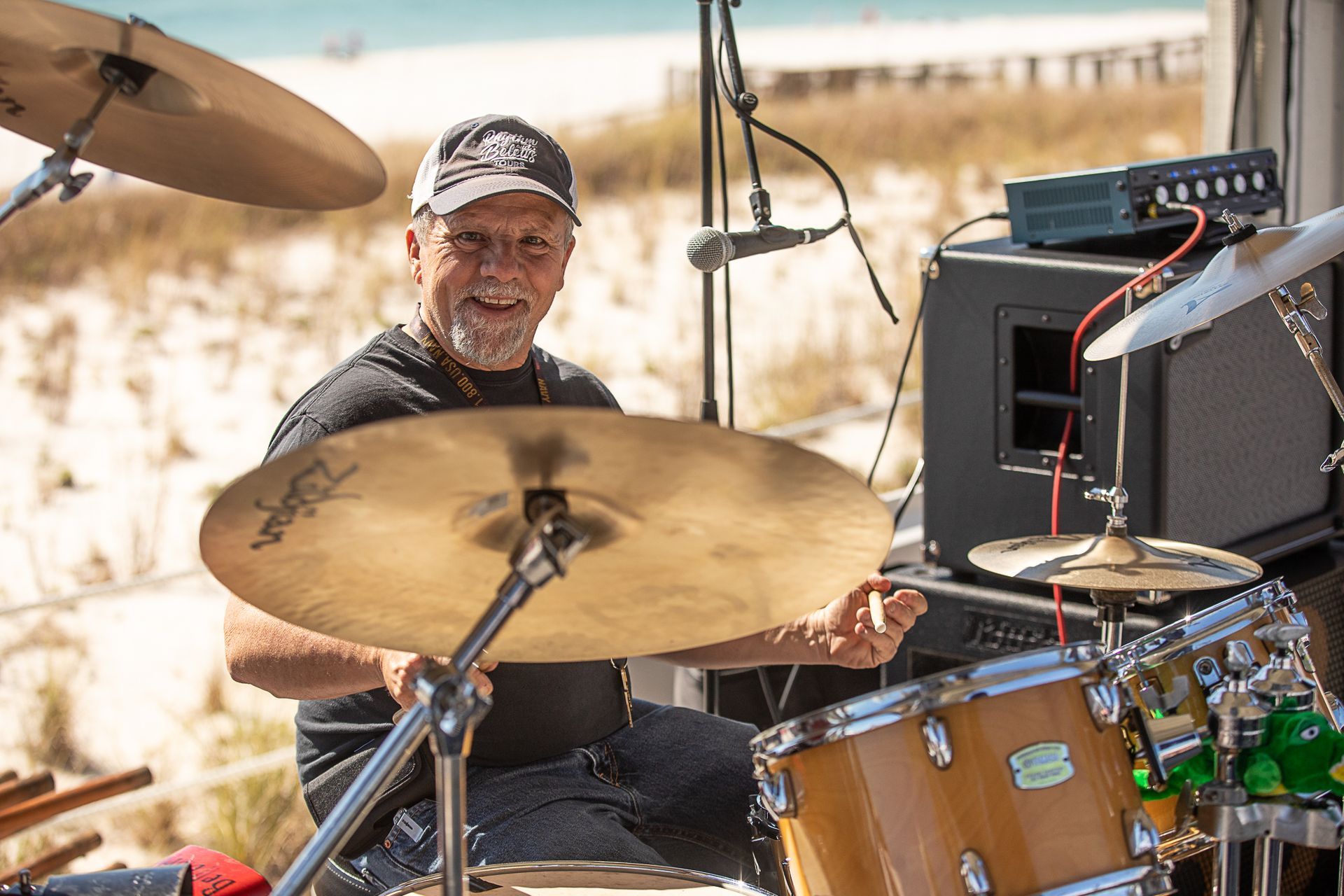 Drummer smiling behind a gold drum kit outdoors in bright sunlight outside of the beach at Coastal Orange Beach