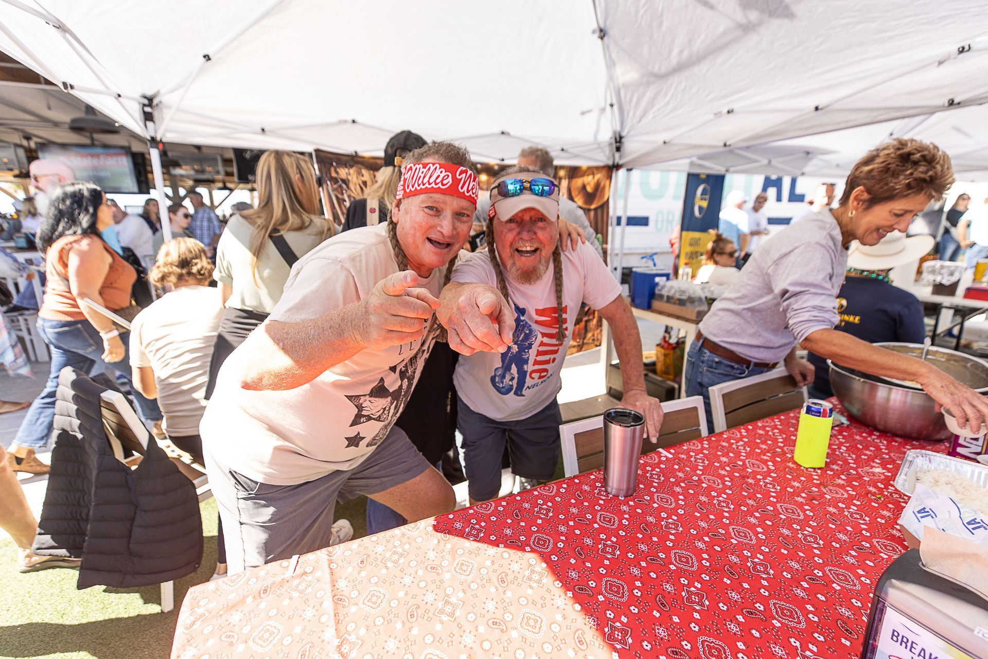 Two smiling volunteers in red bandanas at an outdoor event booth under a tent, serving food on a red tablecloth.