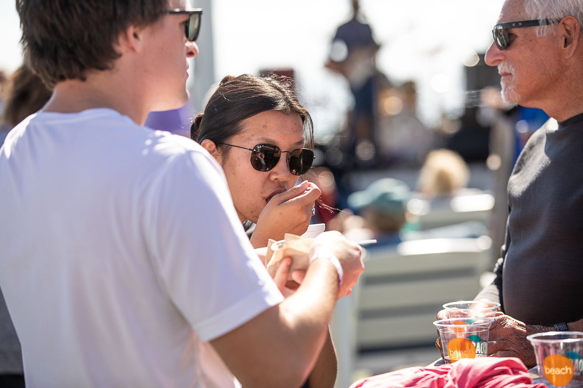 Three people talking at an outdoor table, one holding a drink, in bright sunlight.