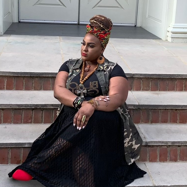 A person wearing a headwrap, camo vest, and black skirt sitting on outdoor steps, looking off to the side.