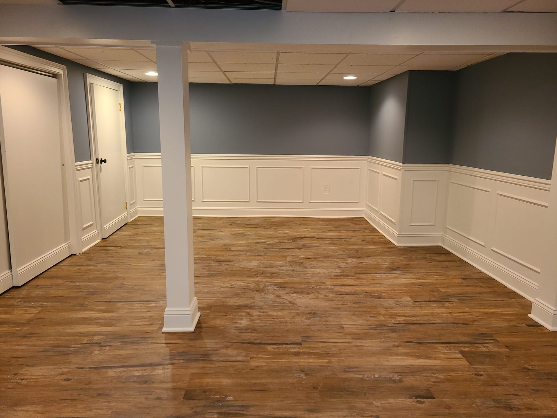 Finished basement interior with wood-look flooring, blue upper walls, white wainscoting, and support columns