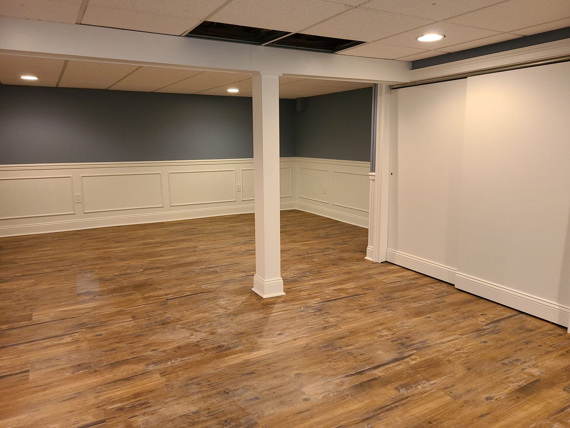 A finished basement with wood-look flooring, white paneling, and gray walls. A central support column is prominent.