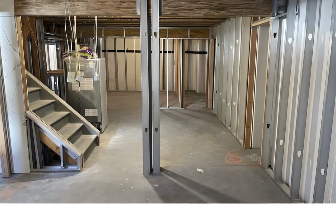 Basement under construction: unfinished walls and stairs, gray and white tones, visible studs and metal framing.