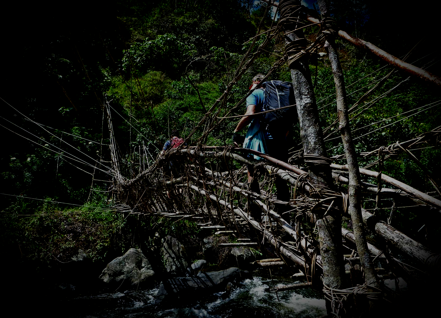 Kokoda Track hikers with backpacks wade through a shallow, rocky stream in a lush green Papua New Guinea jungle.