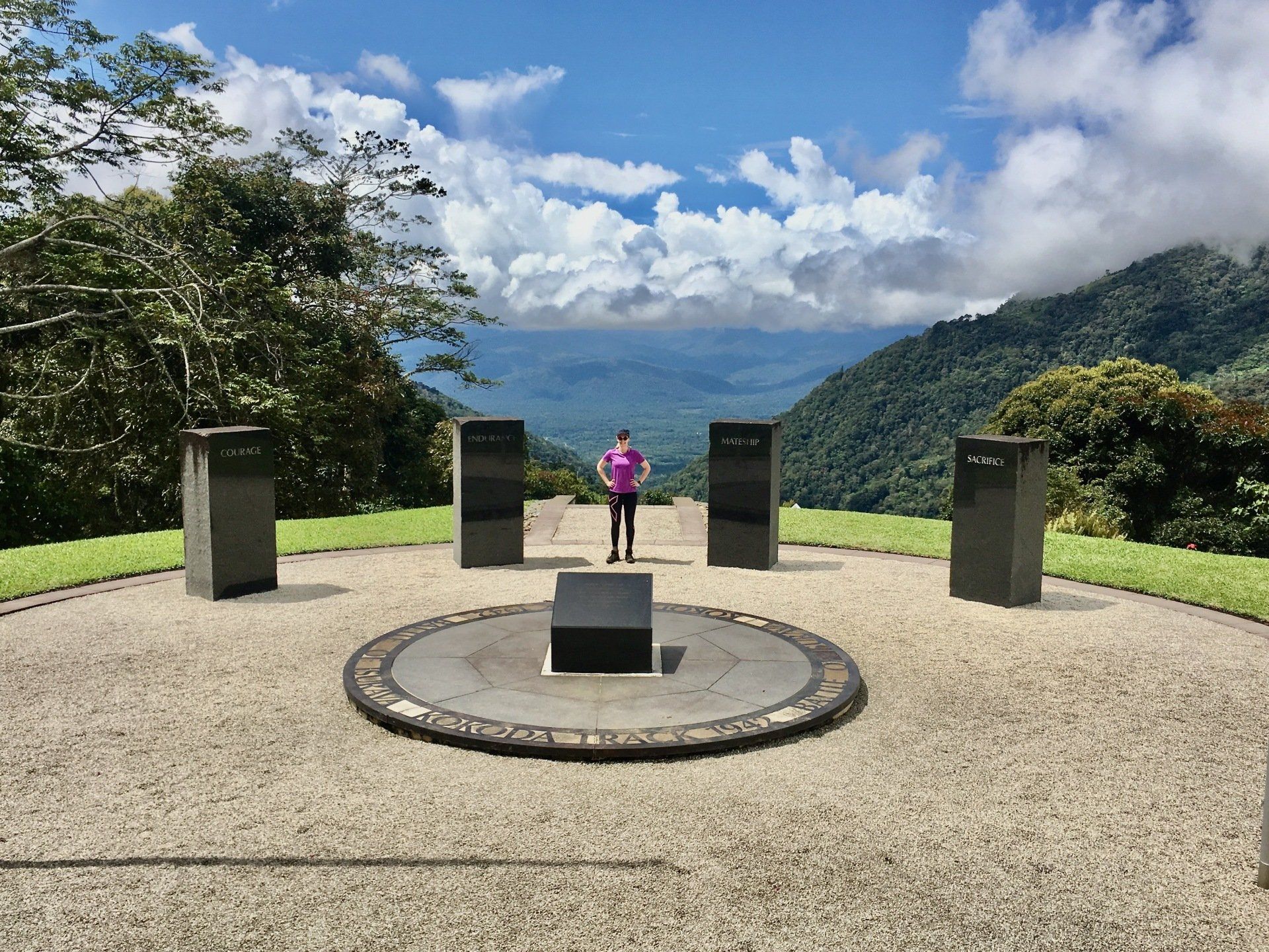 Image depicts the Isurava Memorial at Isurava, Kokoda, PNG. It shows a female standing between the four pillars with the background of the valley. Inscriptions on the four pillars read: Courage, Mateship, Sacrifice and Endurance.