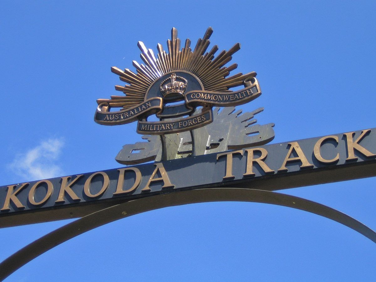 Image depicts the arch at owners corner on the Kokoda Track. Taken from a low position, it faces towards the sky with the words Kokoda Track written on a cross bar and the Australian Commonwealth Military Forces rising sun, crown and inscription on top. Blue sky in the background.