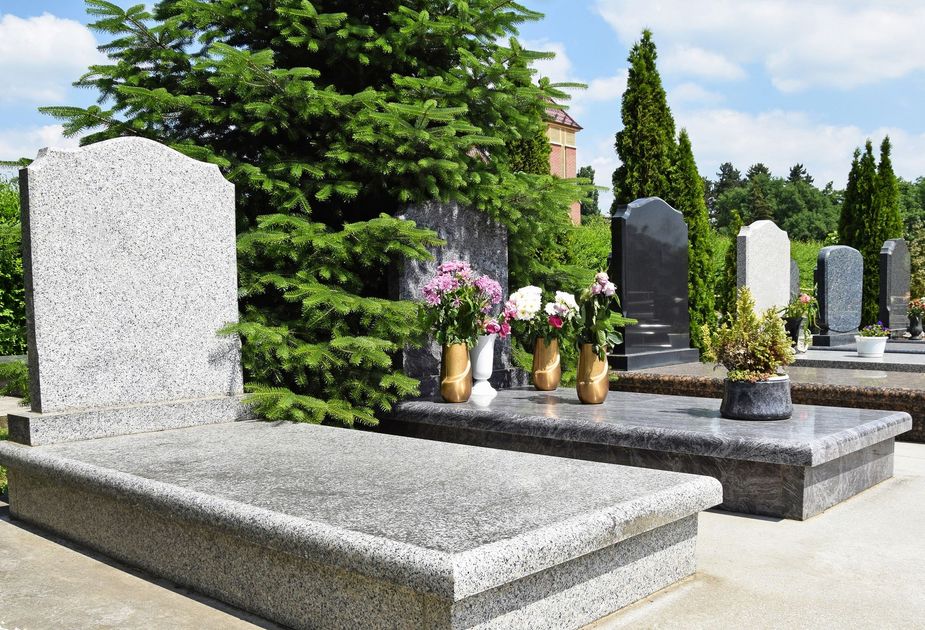 Una fila di lapidi di granito in un cimitero, con fiori accanto a un albero verde sotto un cielo azzurro.