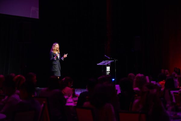 A woman is standing on a stage giving a speech to a crowd of people.
