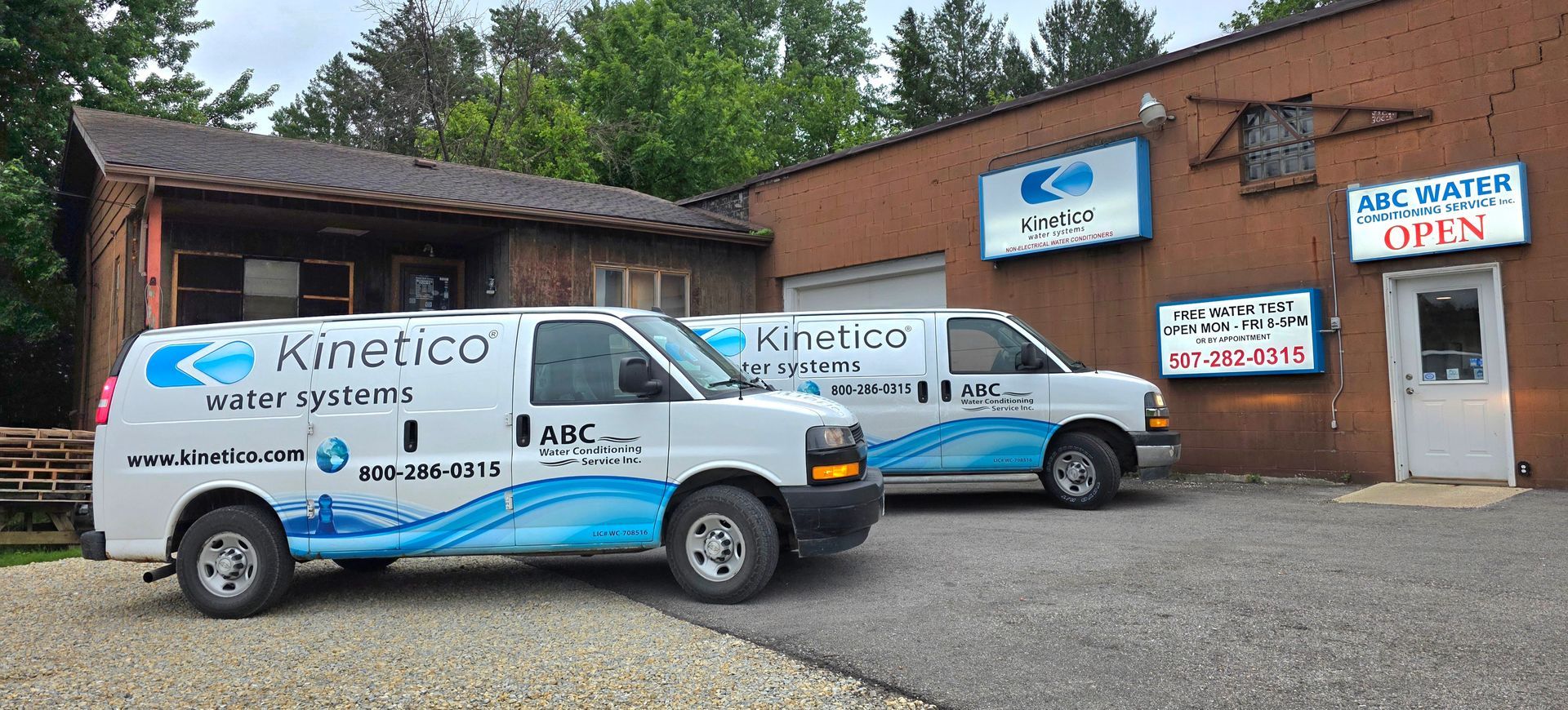 Two Kinetico service vans parked outside a brick building with signs and an open door.