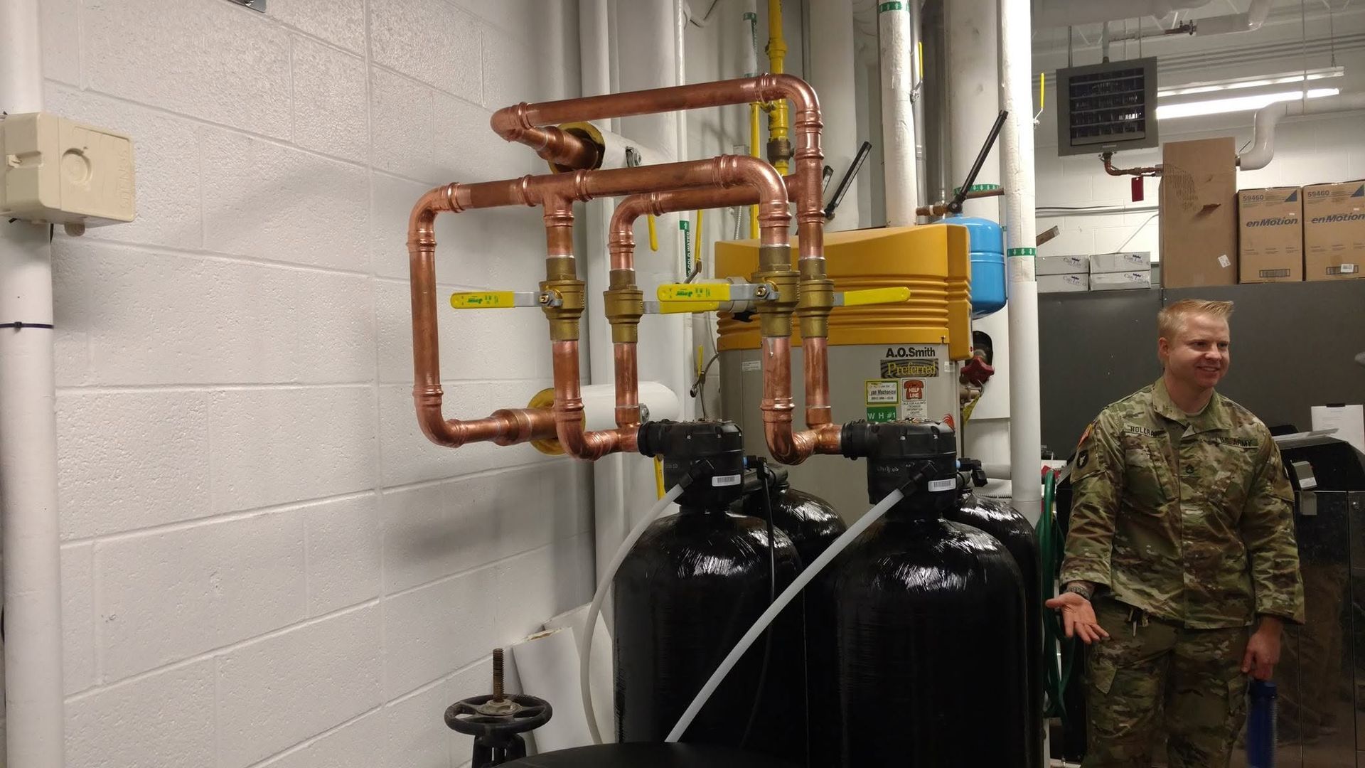 Industrial pipes and tanks in a utility room, with a person in camouflage standing at right.