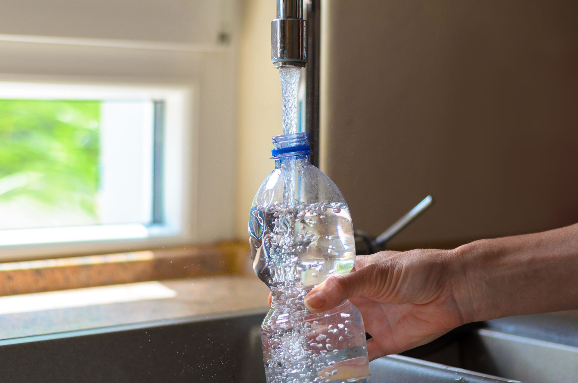 Hand filling a clear plastic bottle with water at a kitchen sink near a window