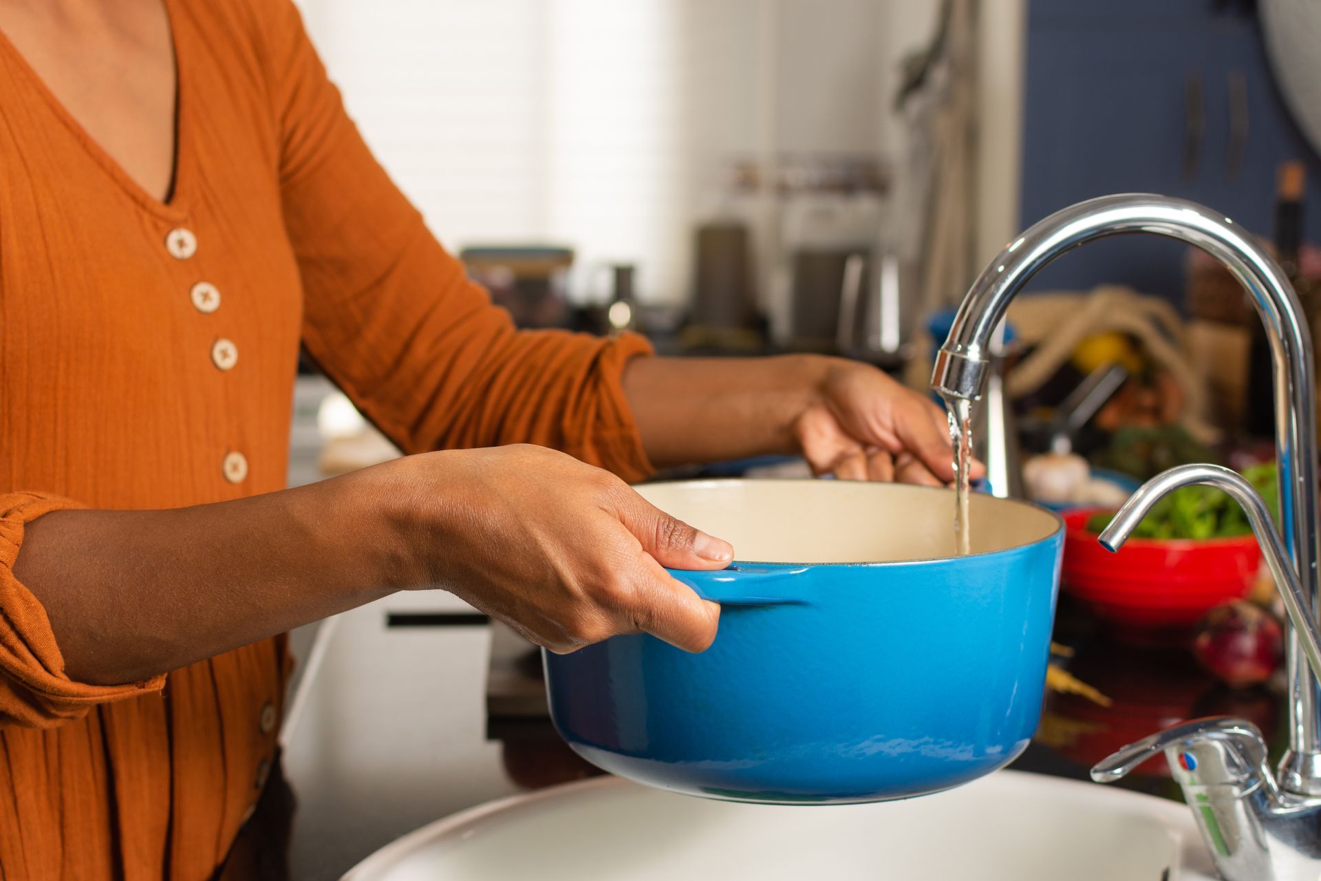 Person washing a blue pot in a kitchen sink while holding a white bowl