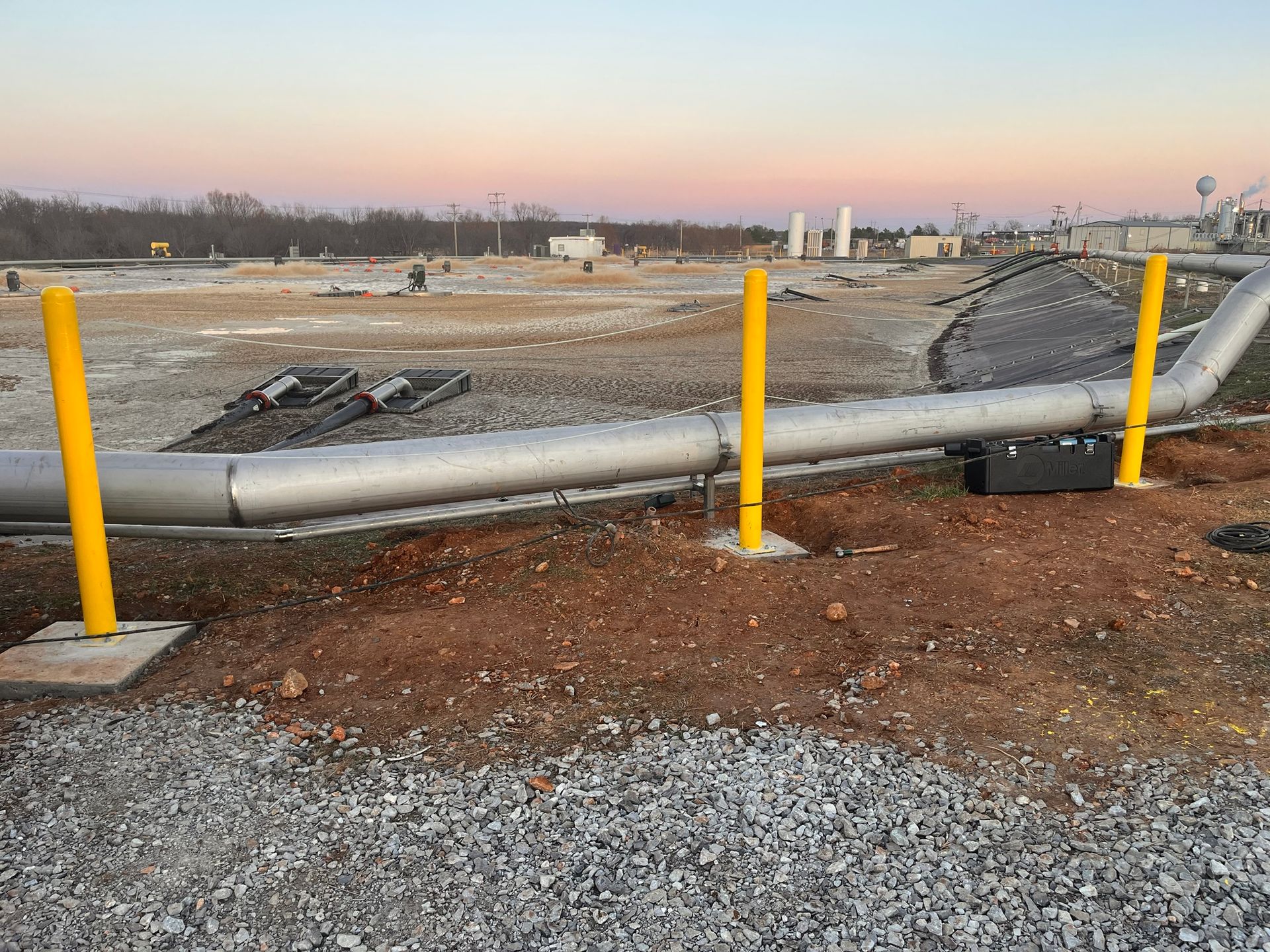 A large pipe is surrounded by yellow poles in a gravel area.