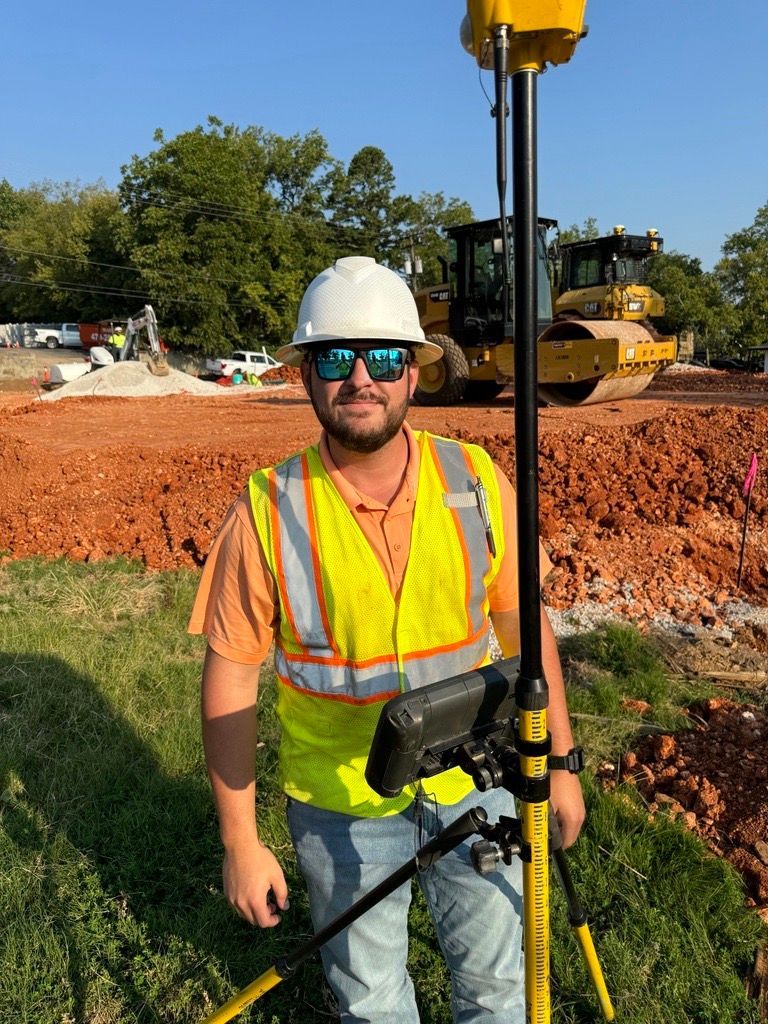 A man in a hard hat and safety vest is standing in a field holding a device.