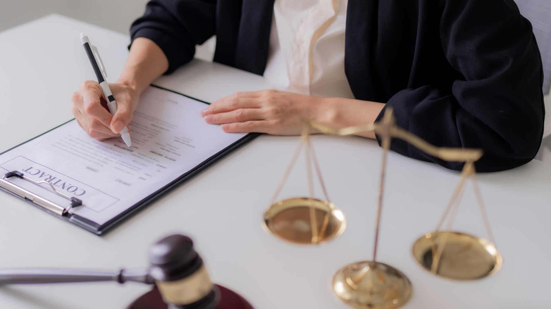 Person writing on contract; scales of justice and gavel on desk.