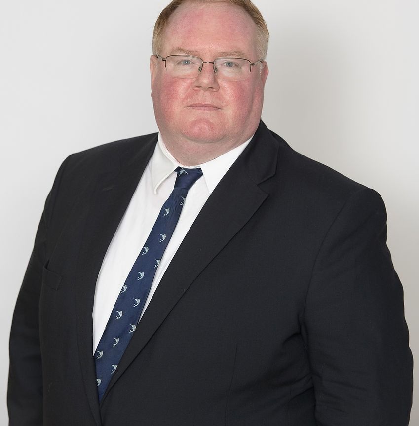 A photo of Brendan J. Dooley in a suit and tie, standing in front of a white wall.