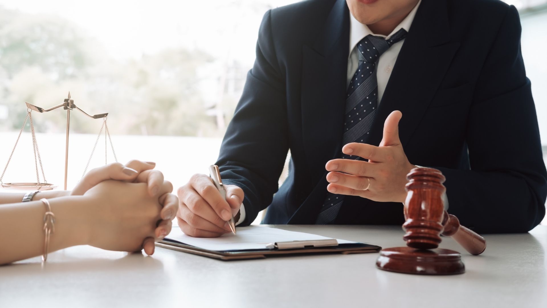 Lawyer in a suit consulting with a person at a desk. Gavel, scales of justice, and documents are present.