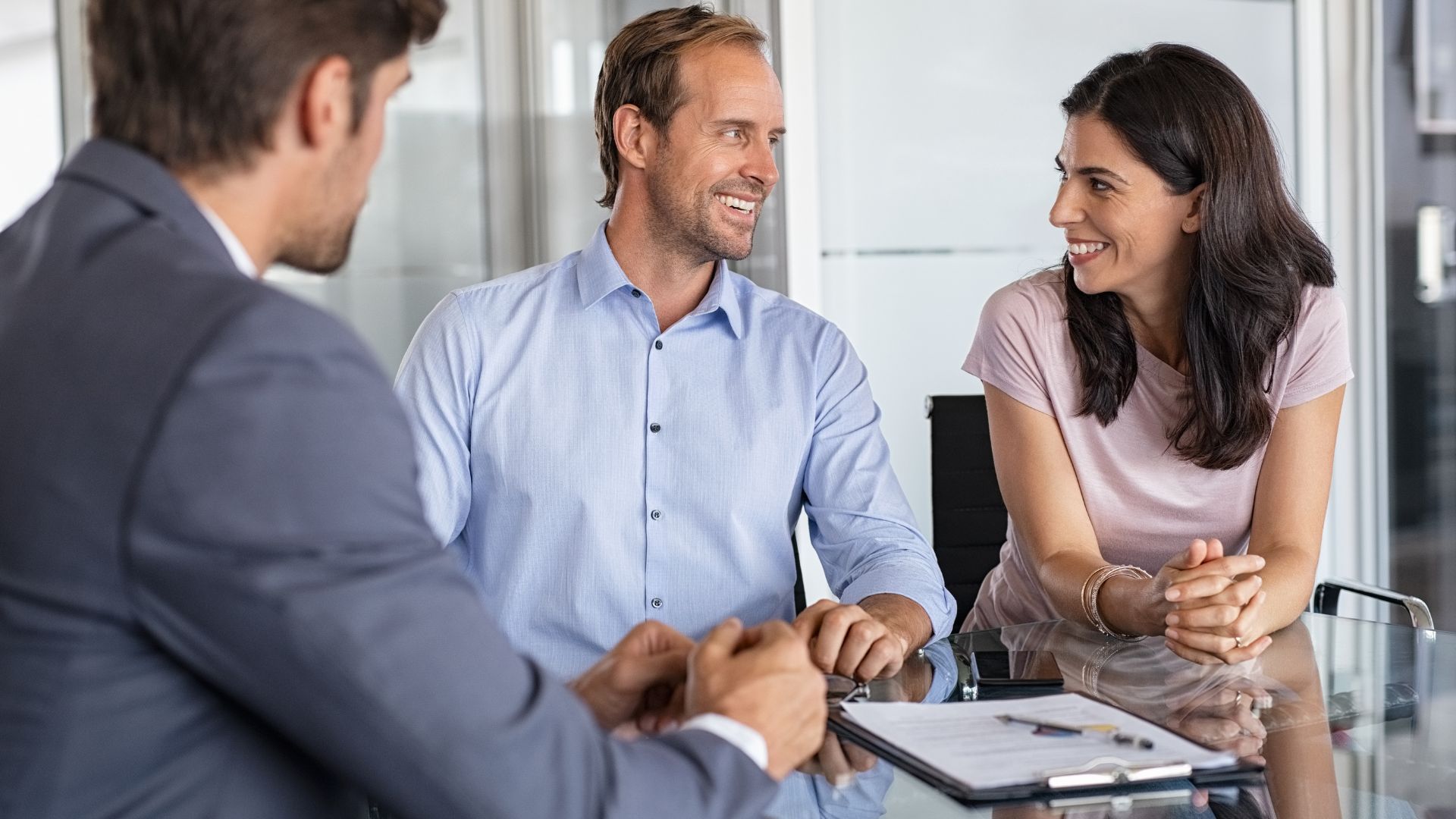 Couple smiling at a person in a suit, sitting at a table with papers, possibly a meeting.
