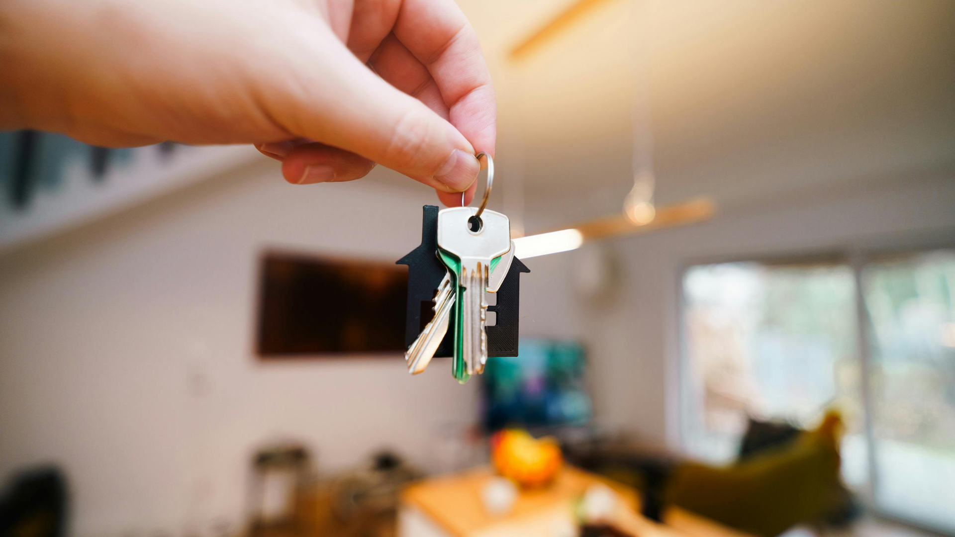 Hand holding house keys in front of a blurred living room interior.