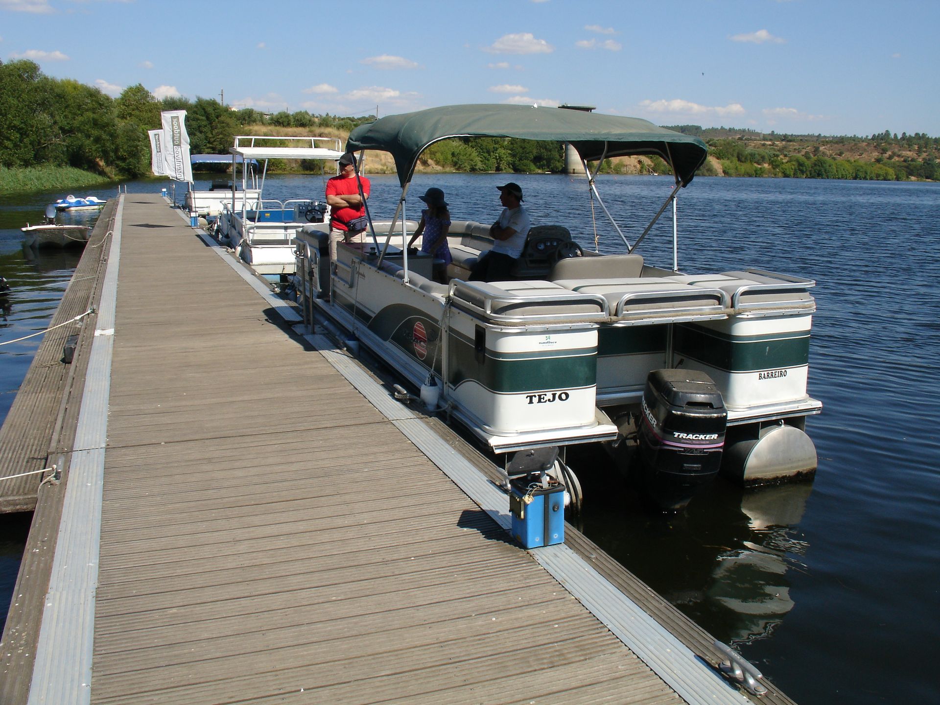 Pontoon boat docked at a lakeside pier with people seated under a canopy on a sunny day