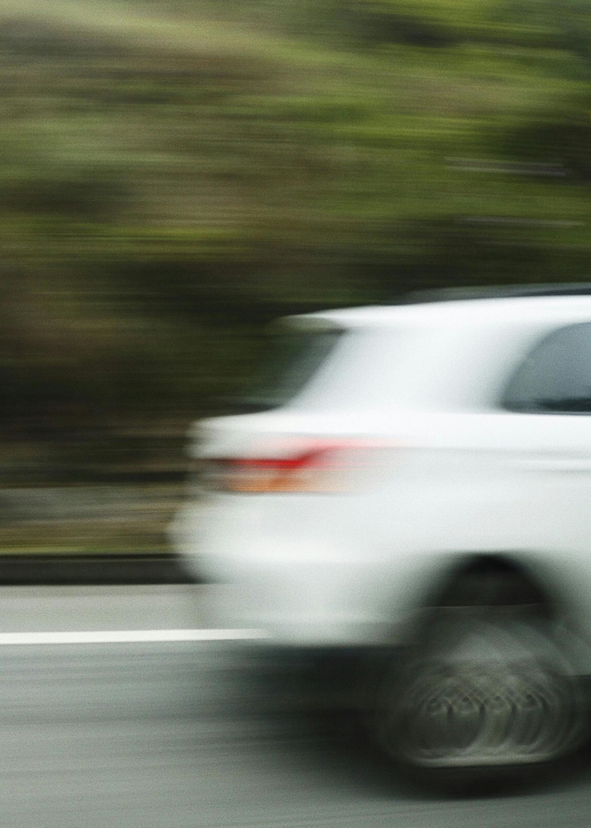 Blurred white car speeding past green roadside trees, creating a motion blur.