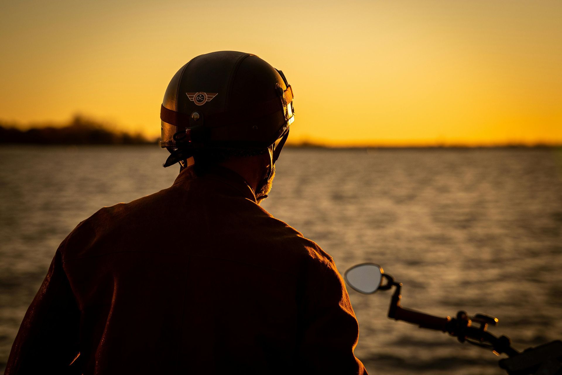 Person on a motorcycle at sunset by the water, silhouetted against a golden sky.