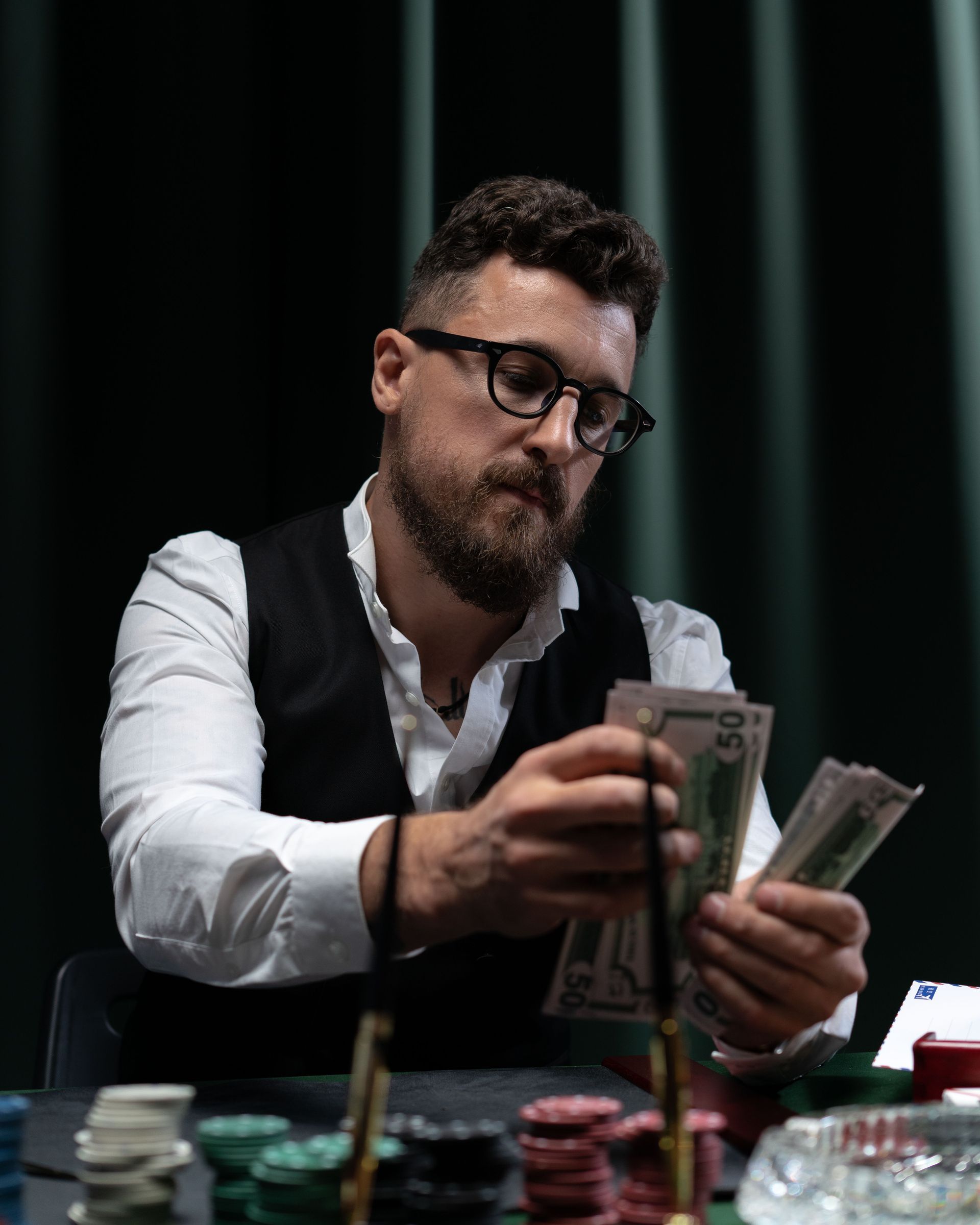 Man in glasses counting cash at a poker table with stacks of chips in a dimly lit room