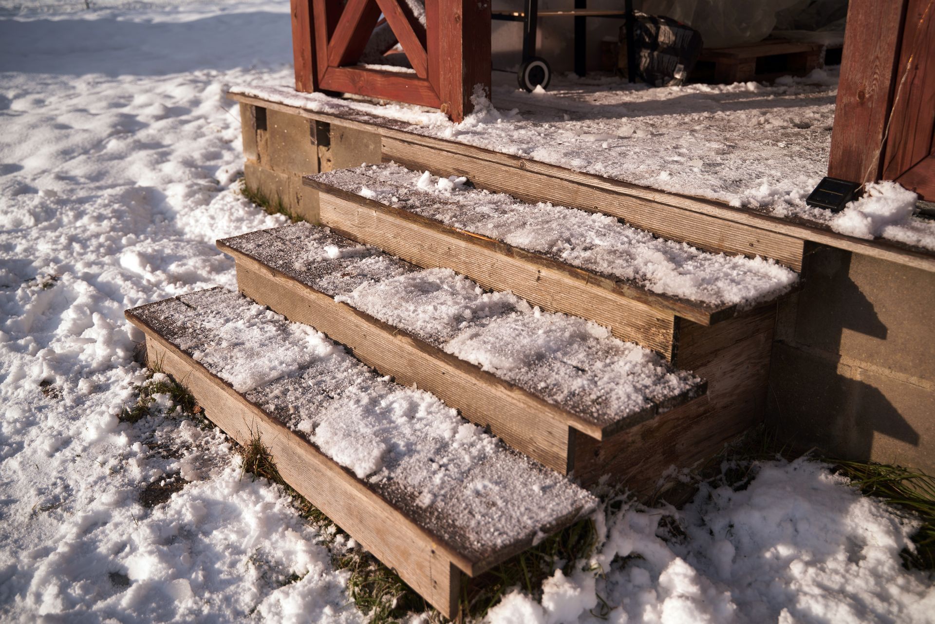 Snow-covered wooden steps leading to a porch beside a red building.