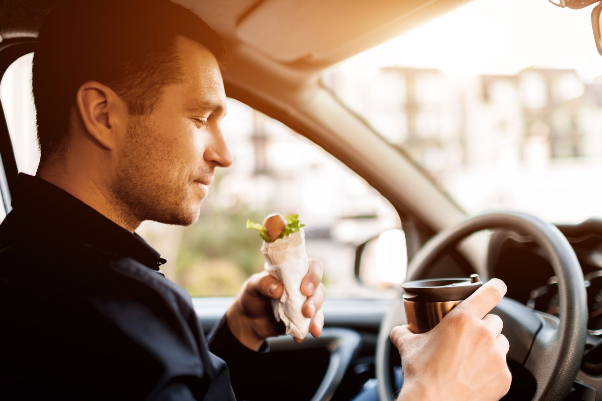 Man drinking coffee and holding a sandwich while driving a car in warm sunlight