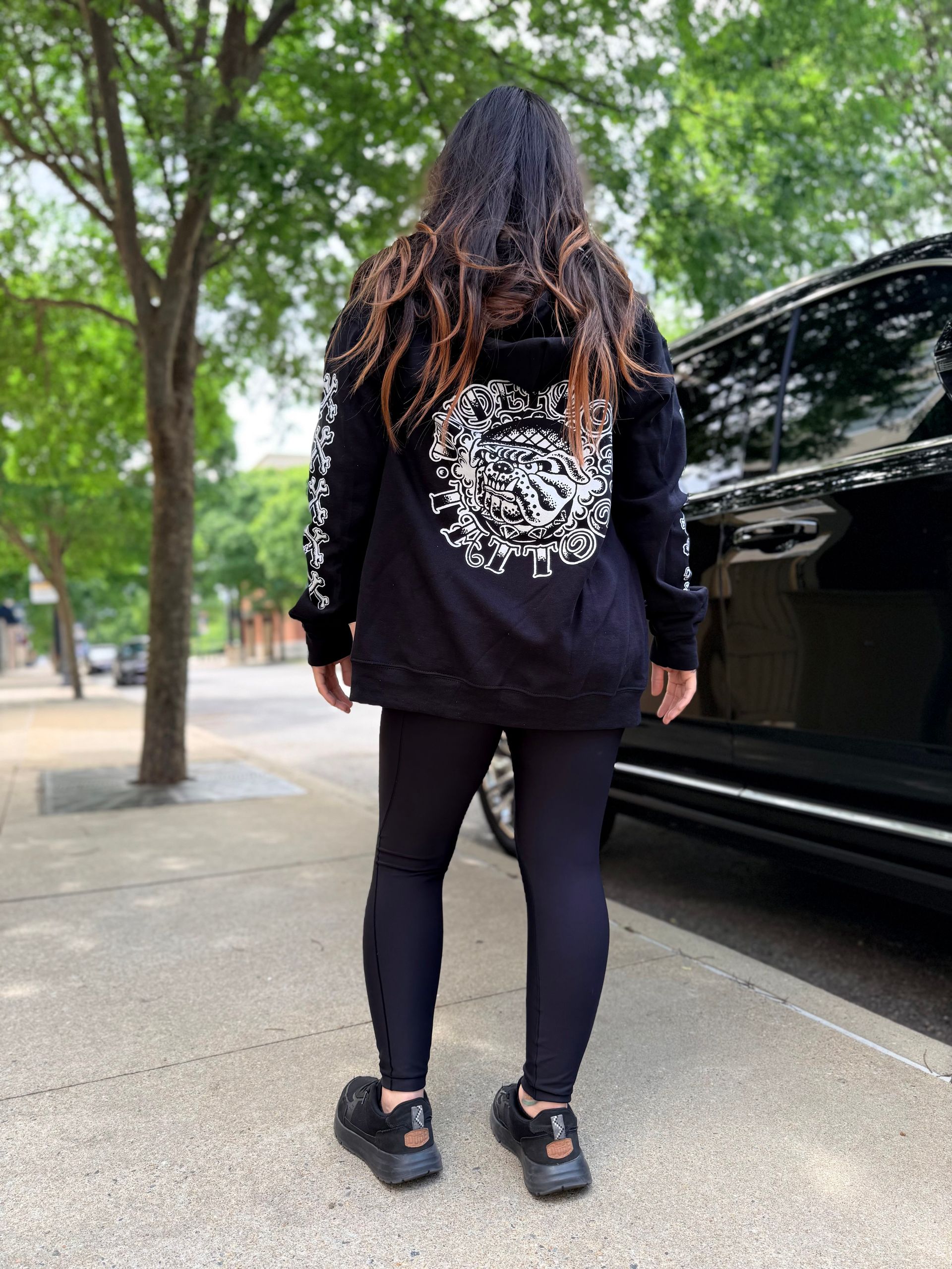 Person in black hoodie and leggings standing on a sidewalk beside a black car, seen from behind.