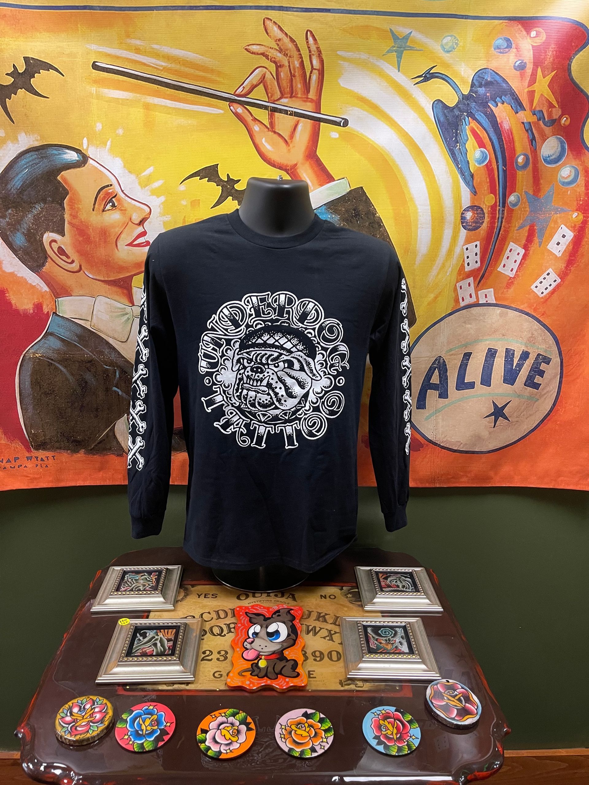 Black long-sleeve shirt on mannequin with white graphic, displayed on a table with colorful pins and art backdrop.