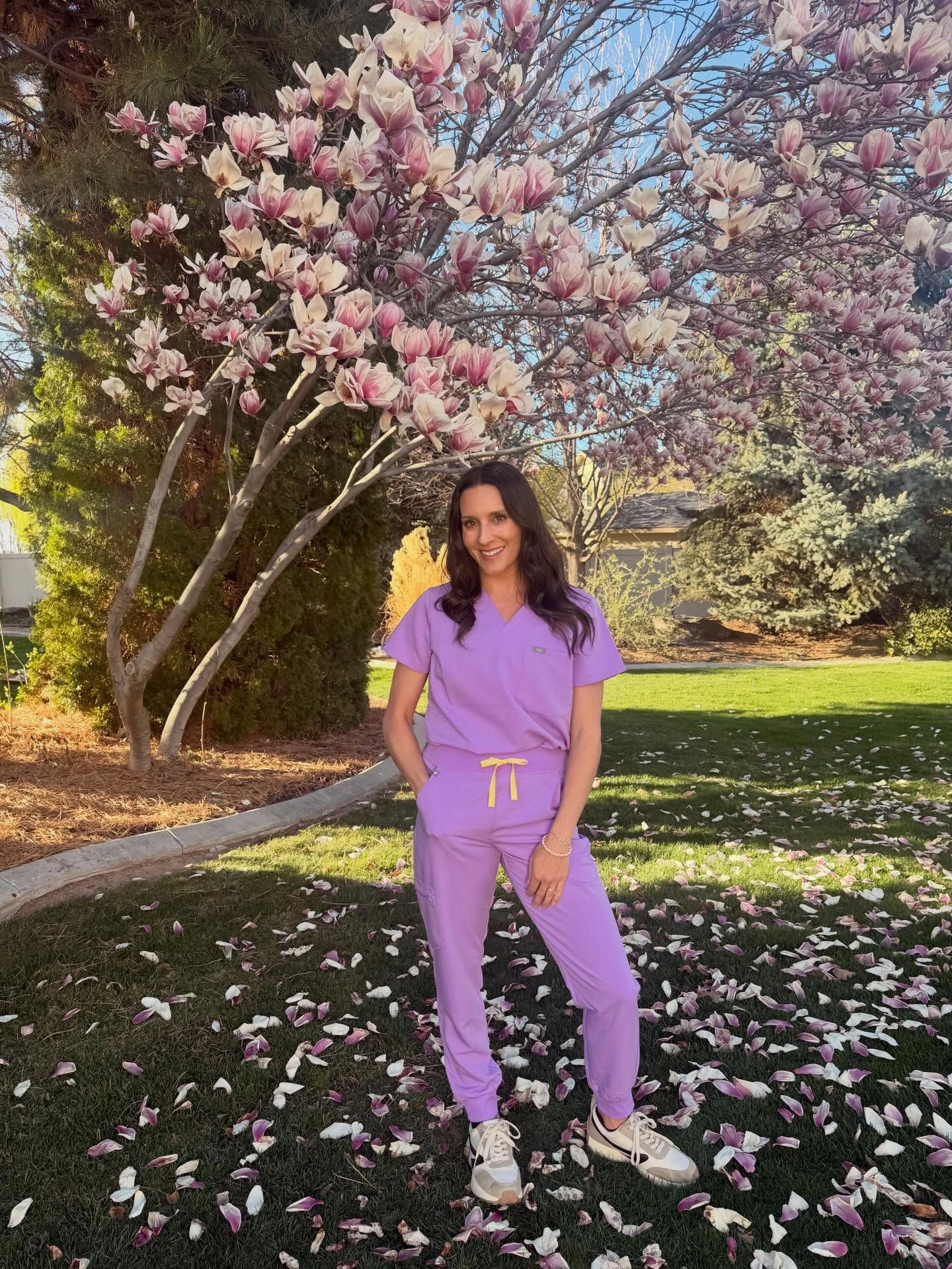Person in purple scrubs standing on a flower-strewn path beneath blooming trees.
