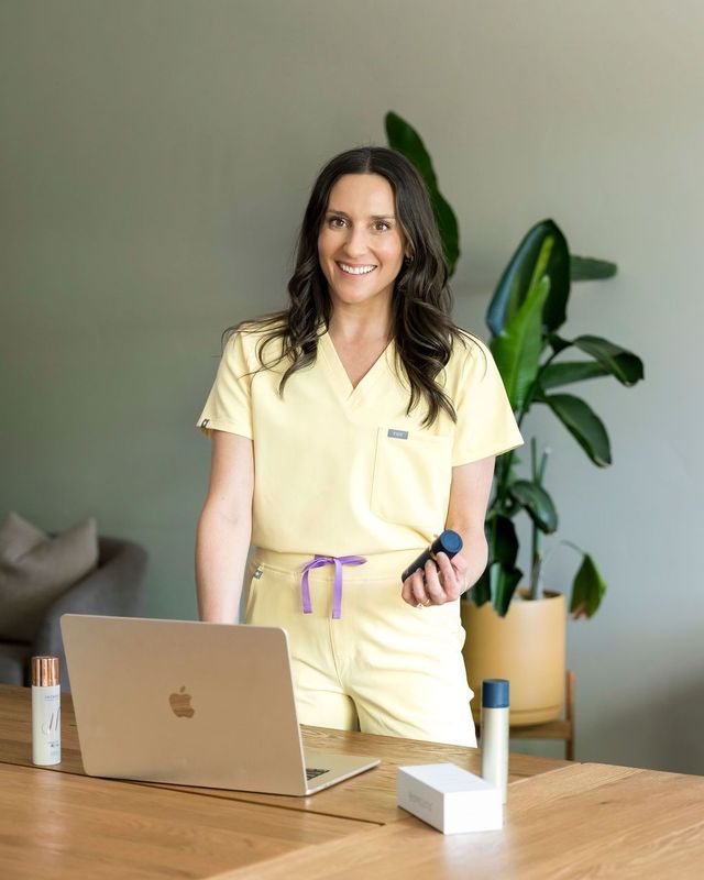 Woman standing beside a desk with a laptop and plants, smiling in a home office.