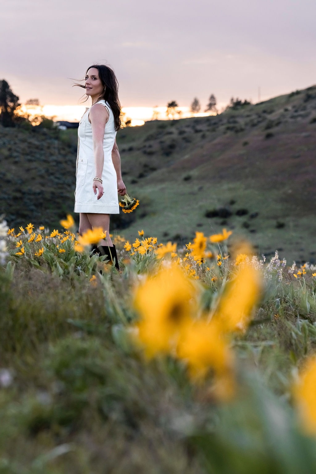 Woman in a white dress standing in a field of yellow flowers at sunset with mountains in the background