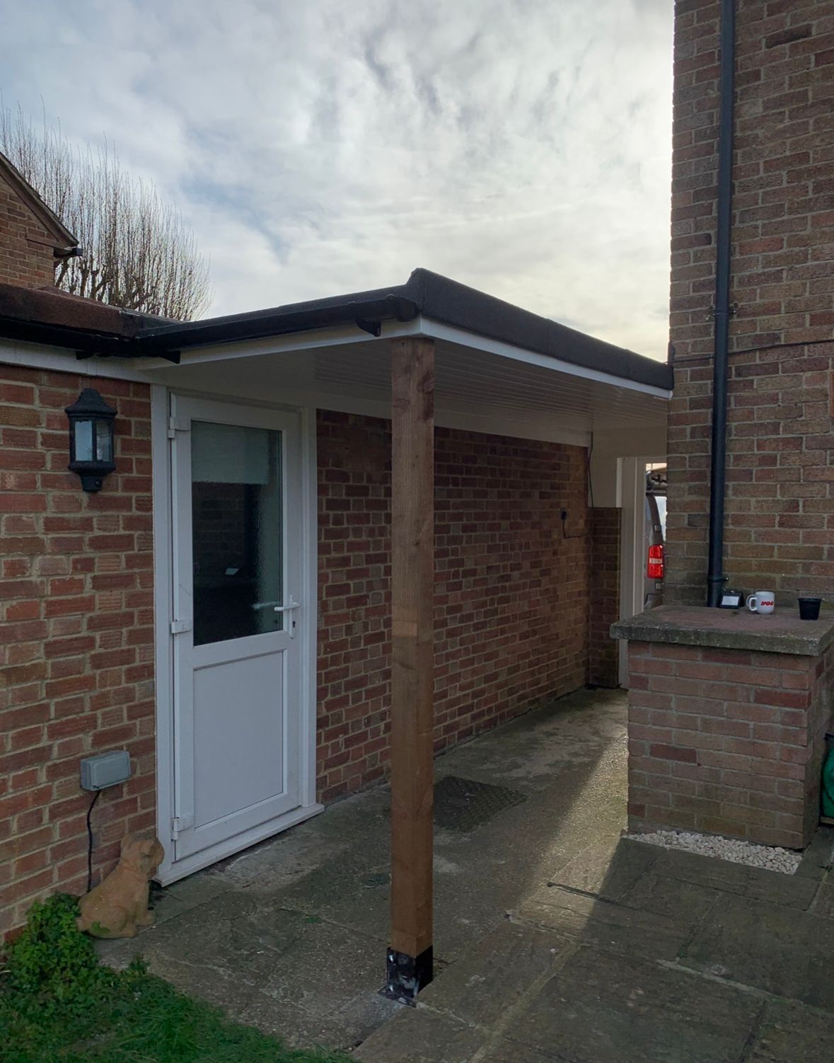 Covered walkway with brick walls and a white door, supported by a wooden column under a gray sky.